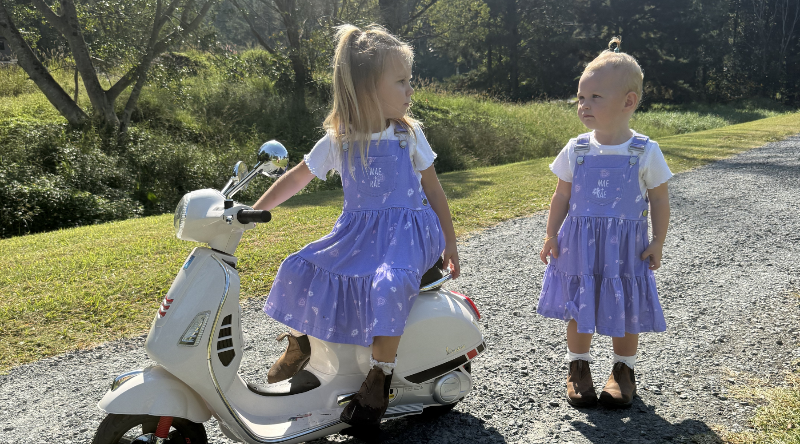 Two children in matching daydream dresses from mae and rae standing next to a scooter on a path.