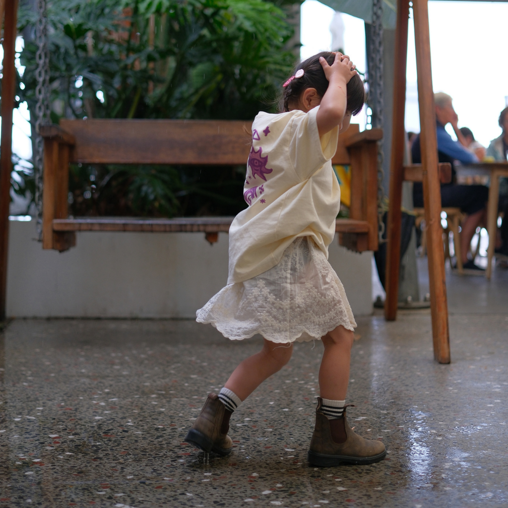 Toddler walking in the Butter Cosmic Tee and a lace skirt, showing the purple back print