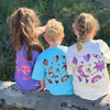 Three kids on a log wearing Cosmic Tees in Grape, Aqua and Butter, showing the moon, sun and star back print