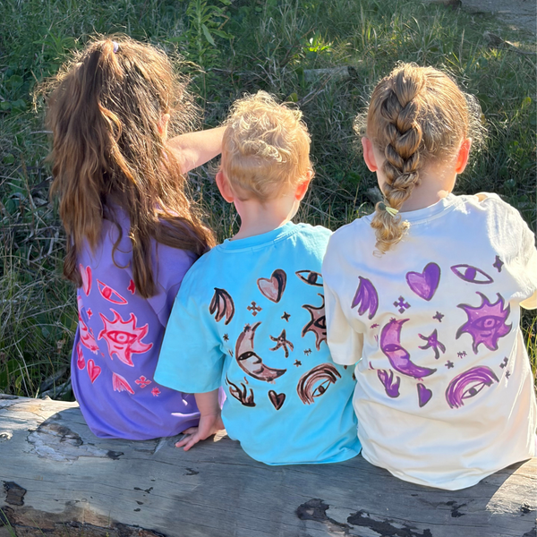 Three kids on a log wearing Cosmic Tees in Grape, Aqua and Butter, showing the moon, sun and star back print