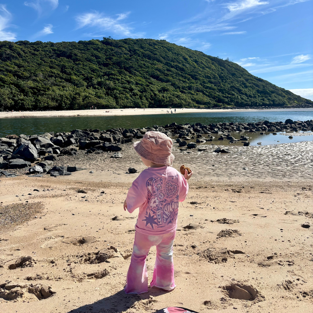 Toddler at the beach in a bucket hat wearing the Baby Pink Dreamers Tee and pink Stardust Flares